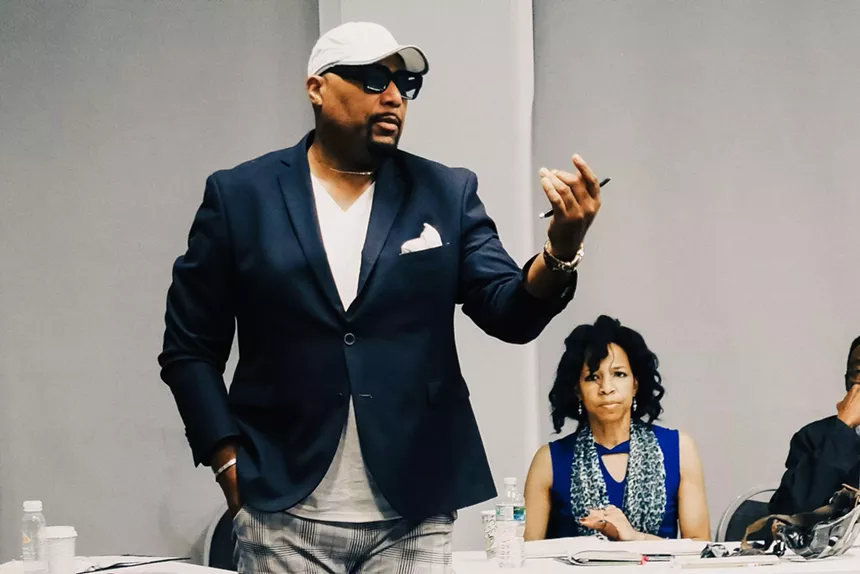 Layon Gray standing with his arm extended while Carey Brianna Hart is seated behind him