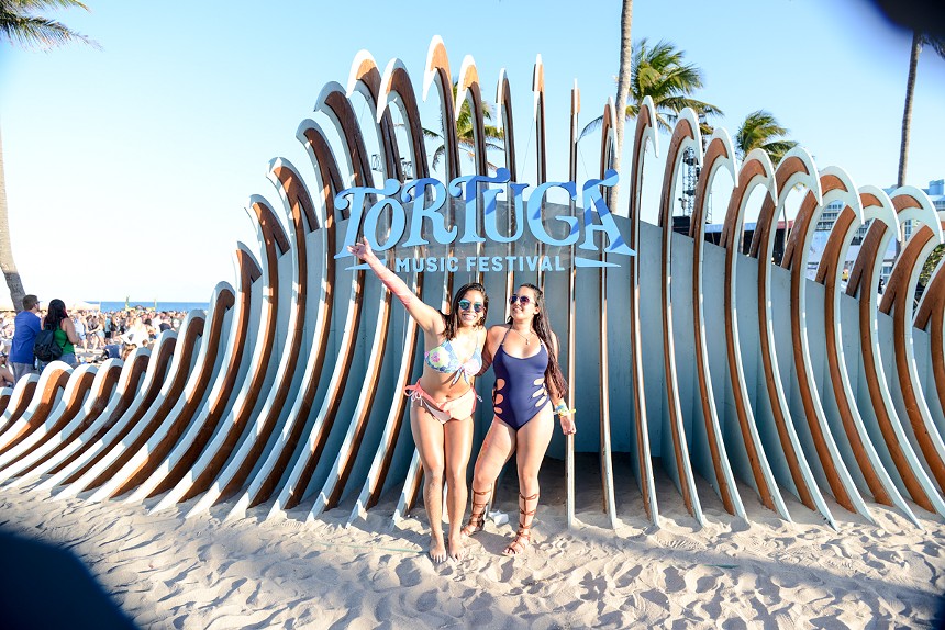 Two women pose in front of the logo for Tortuga Music Festival
