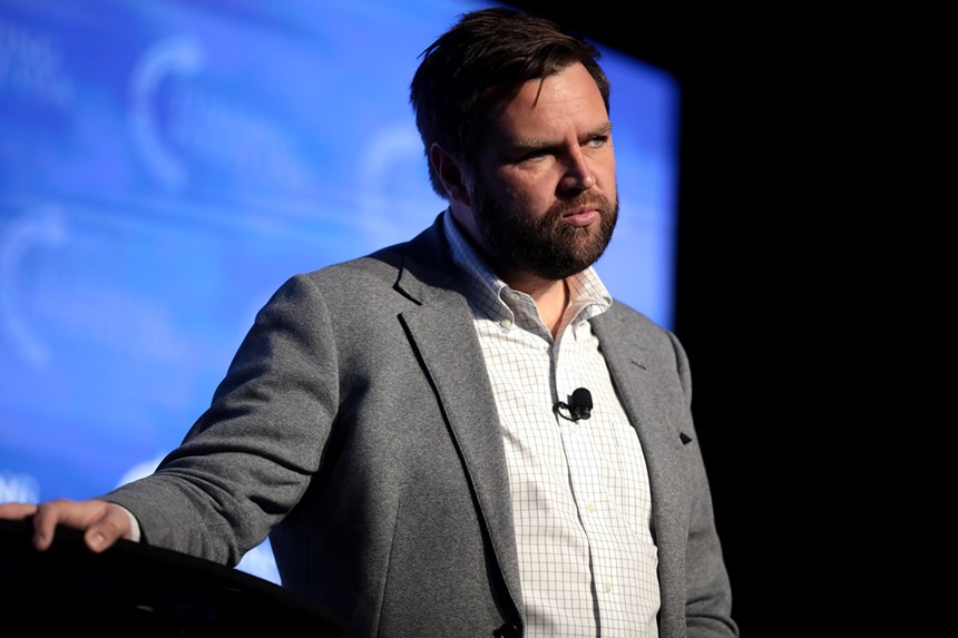 J.D. Vance wearing jacket with no tie and open collar, with right hand resting on a podium, looking serious