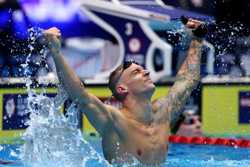 Swimmer Caeleb Dressel lifts his arms of a pool in a gesture of victory