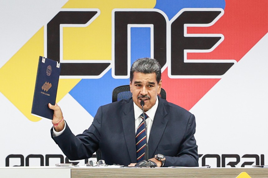 Nicolas Maduro, dressed in a suit, holds up a blue folder with an election certificate
