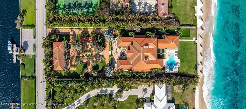 Aerial shot of the mansion showing tennis court, palm-lined driveway, and water access