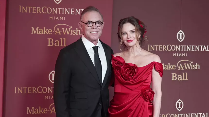 Shareef M. Malnik and Gabrielle Anwar pose on the red carpet. Anwar wears a red, floral-embellished gown, while Malnik is in a classic black tuxedo.