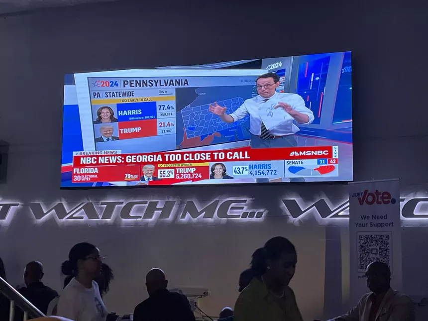 TV screen showing election results with attendees watching at Just Vote Period watch party