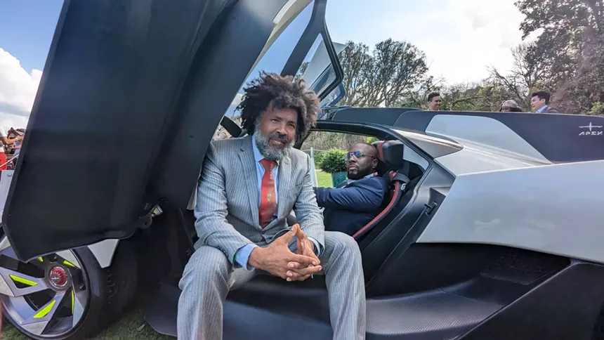 Elo King sits at the open door of a sleek Attucks Apex AP0 supercar, wearing a gray pinstripe suit and red tie, while Wyclef Jean lounges in the driver's seat, dressed in a navy suit, during the 2023 Amelia Concours d'Elegance.