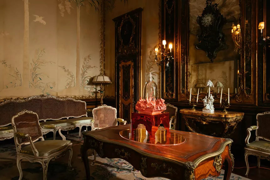 A sculpture made of glass and ceramics sits atop a wooden table surrounded by chairs, candelabras, and other decor.