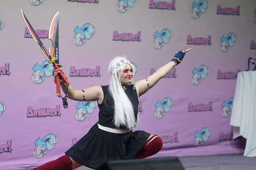 a person in a long blonde wig and costume poses in front of a step and repeat banner reading "Animate!"