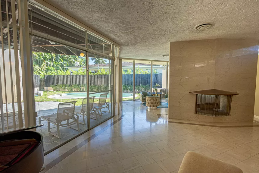 A tiled living room with a double-sided fireplace looks out to a curved pool