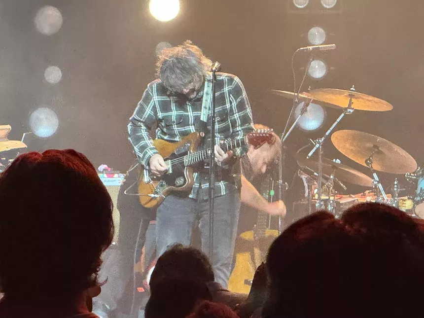 Wilco frontman Jeff Tweedy on stage, rocking, with the silhouettes of audience members in the foreground