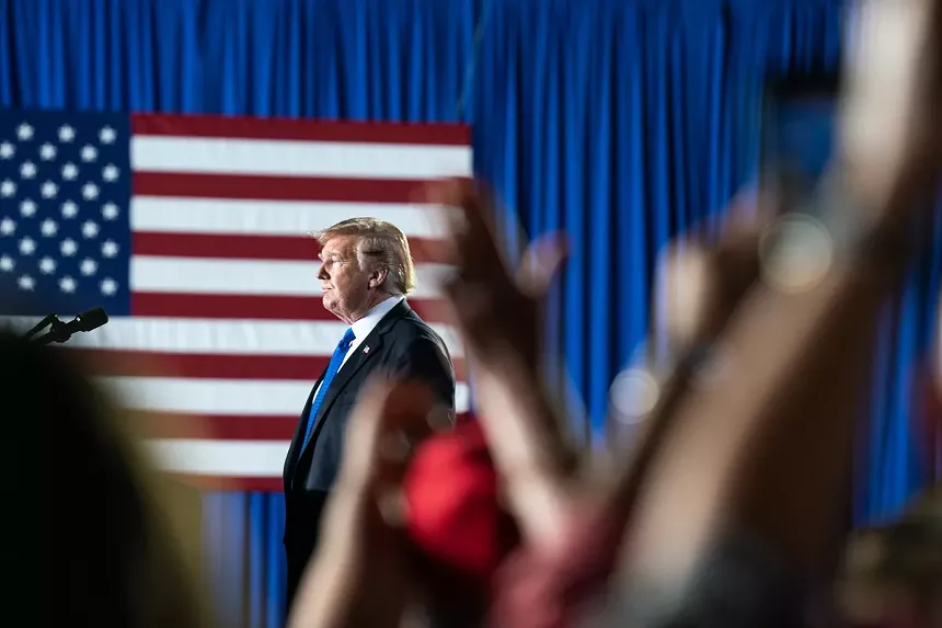 trump in profile, with an american flag in the background and supporters' raised arms in the foreground