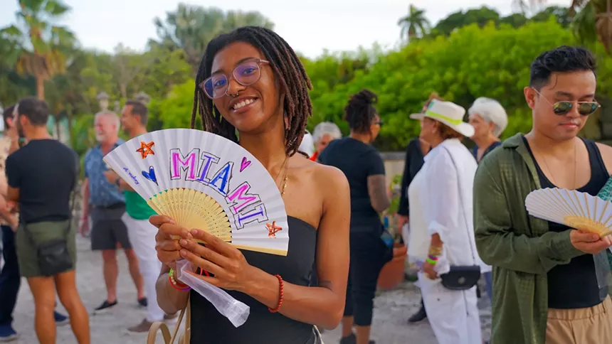 a person holds a handmade fan reading "Miami" at a Pride Month event