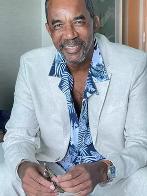 photo of a smiling, goateed black man wearing a cement gray sport coat over a blue tropical print shirt and holding a pair of sunglasses