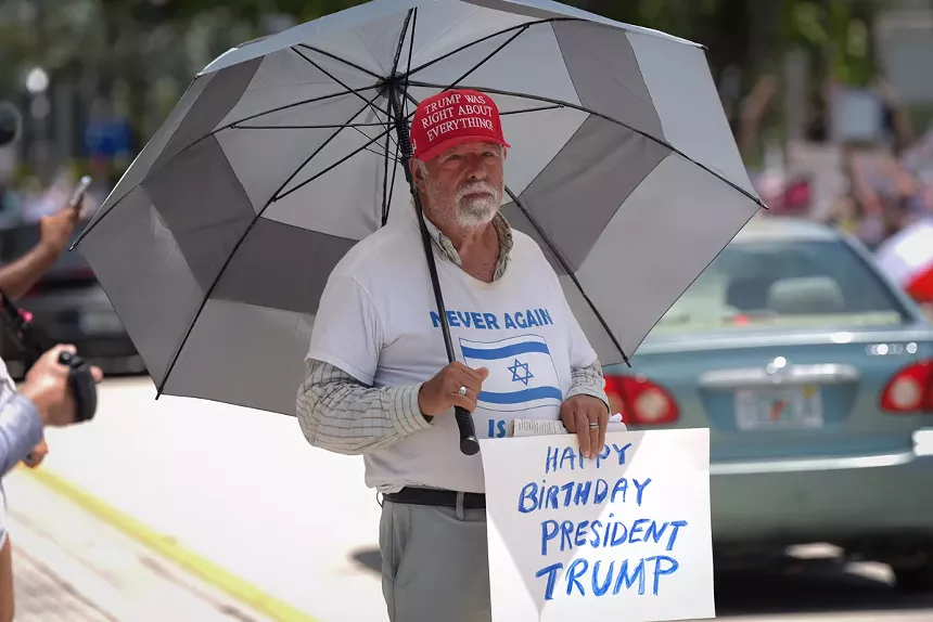 A man wearing a red cap reading, "Trump was right about everything," and a shirt with an Israeli flag and partially obstructed text holds a protest sign reading, "Happy birthday, President Trump"