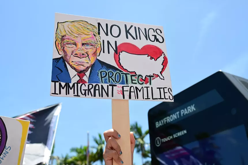 A home-made sign depicts a map of the U.S. inside a heart, a drawing of Donald Trump's face with a pig's nose, and text reading, "No kings in...Protect immigrant families."