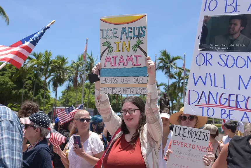 A woman holds up a homemade sign reading, "ICE melts in Miami. Hands off our friends & neighbors."