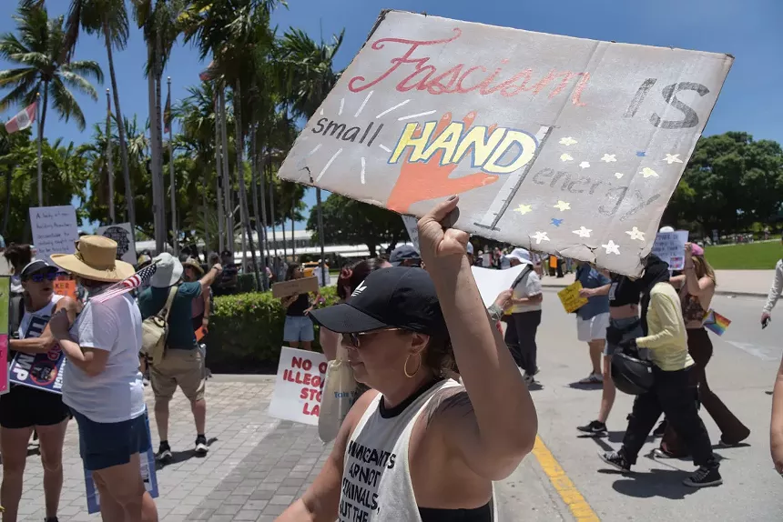 A woman holds a sign reading, "Fascism is small hand energy" with a painting of an orange hand and a ruler