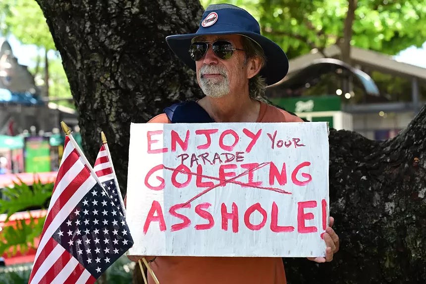 Man holds up a sign reading, "Enjoy golfing asshole!" The word golfing is crossed out and replaced with "Your parade"