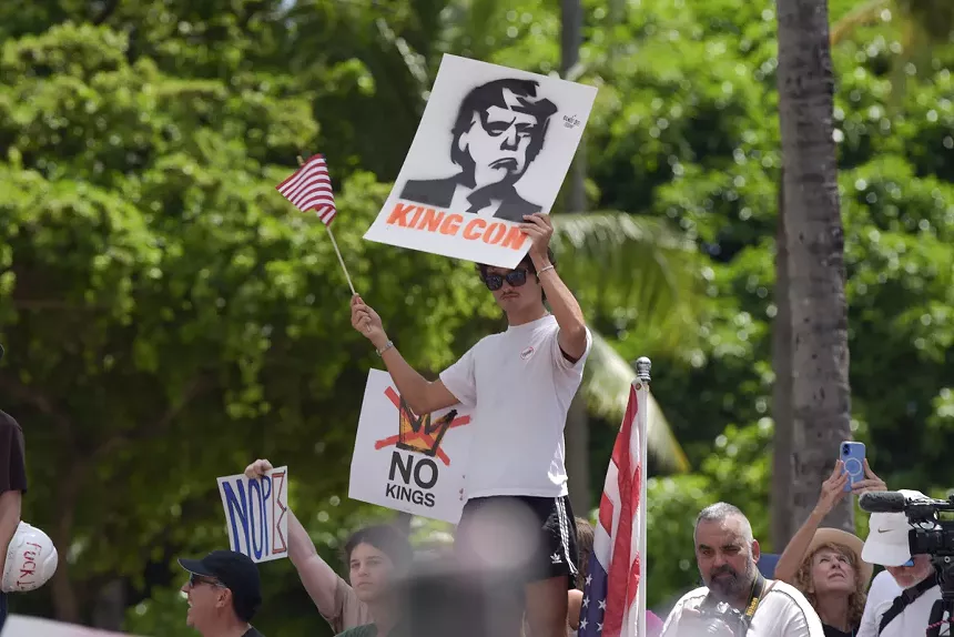 A protester holds up a stencil of Donald Trump's face with text reading, "King Con."