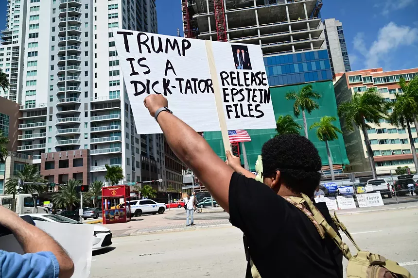 A man holds up a sign reading, "Trump is a dick-tator" and "Release the Epstein Files"