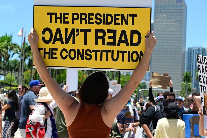 protester holding up an all-caps yellow sign that says "The president can't read the Constitution" in black type with the words "Can't Read" rendered in letters that alternate between backward and forward