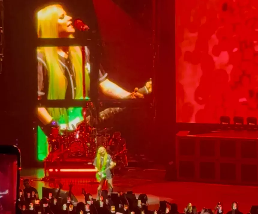 Singer Avril Lavigne plays a green guitar onstage. A Jumbotron behind her projects her image.