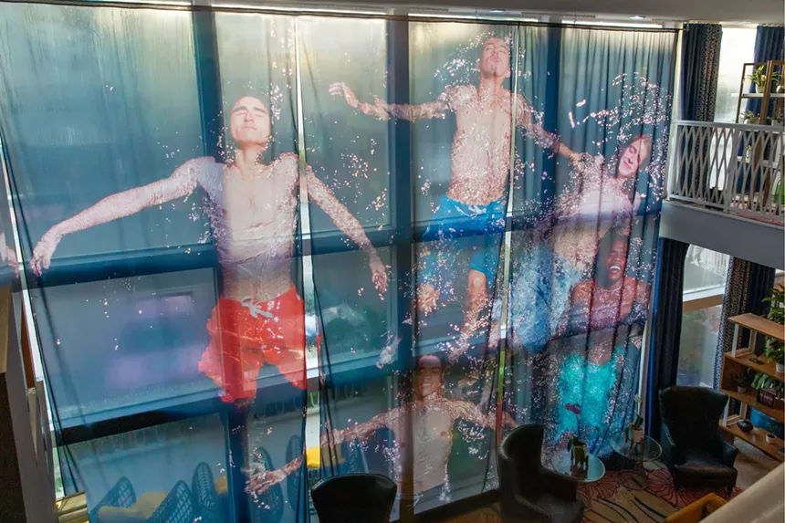 A photograph printed on fabric hangs in front of floor-to-ceiling windows at a hotel. The photograph depicts five young men swimming