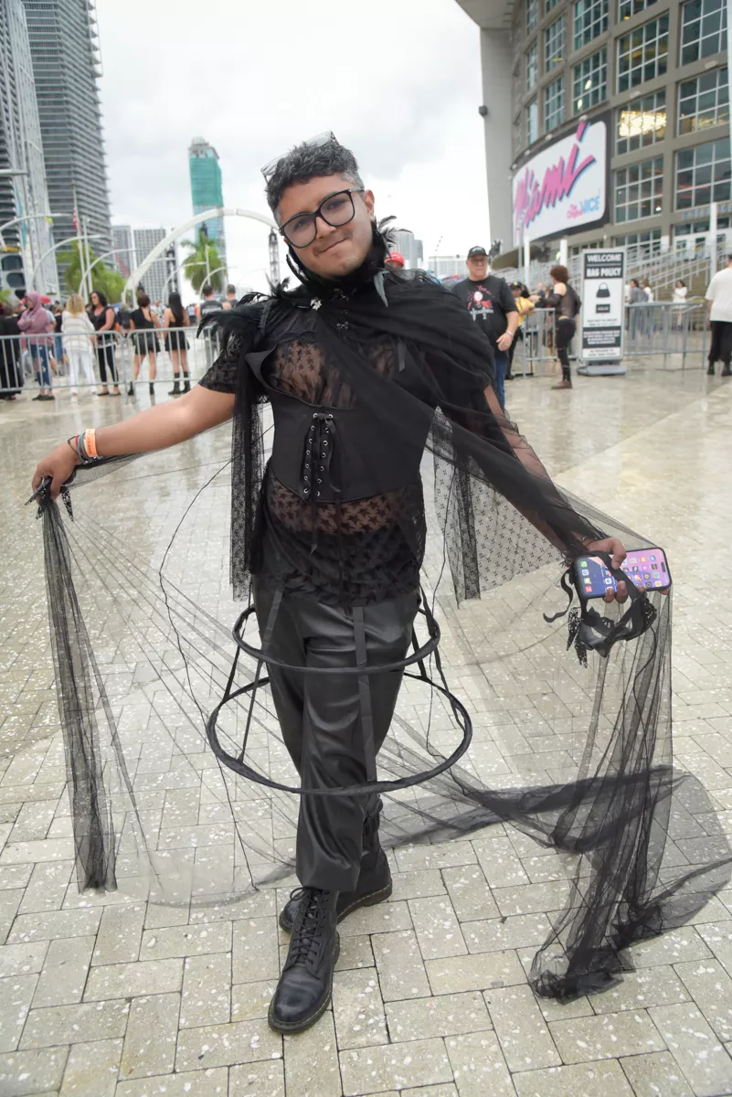 A Lady Gaga fan poses in front of an arena, showing off their outfit: a feathery black, high collar, corset, and tulle cape
