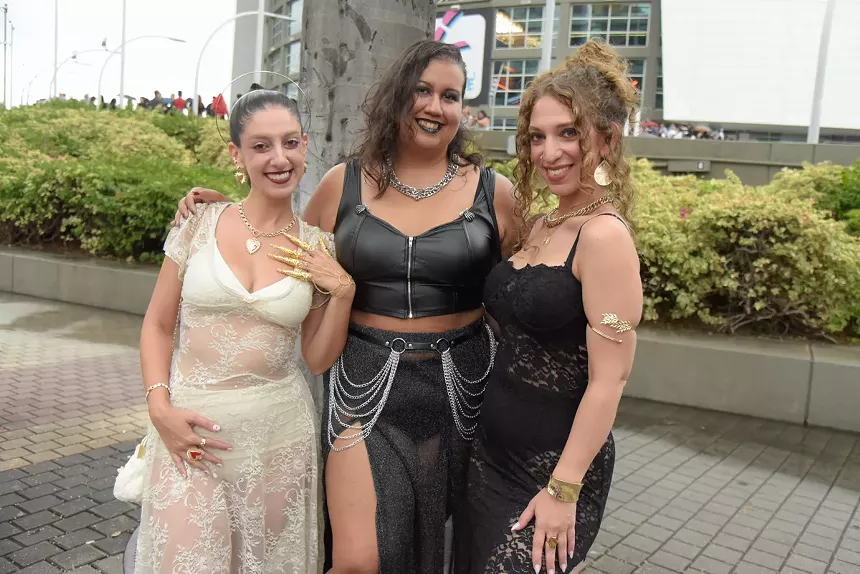 Three women pose in lace and leather outfits in front of an arena