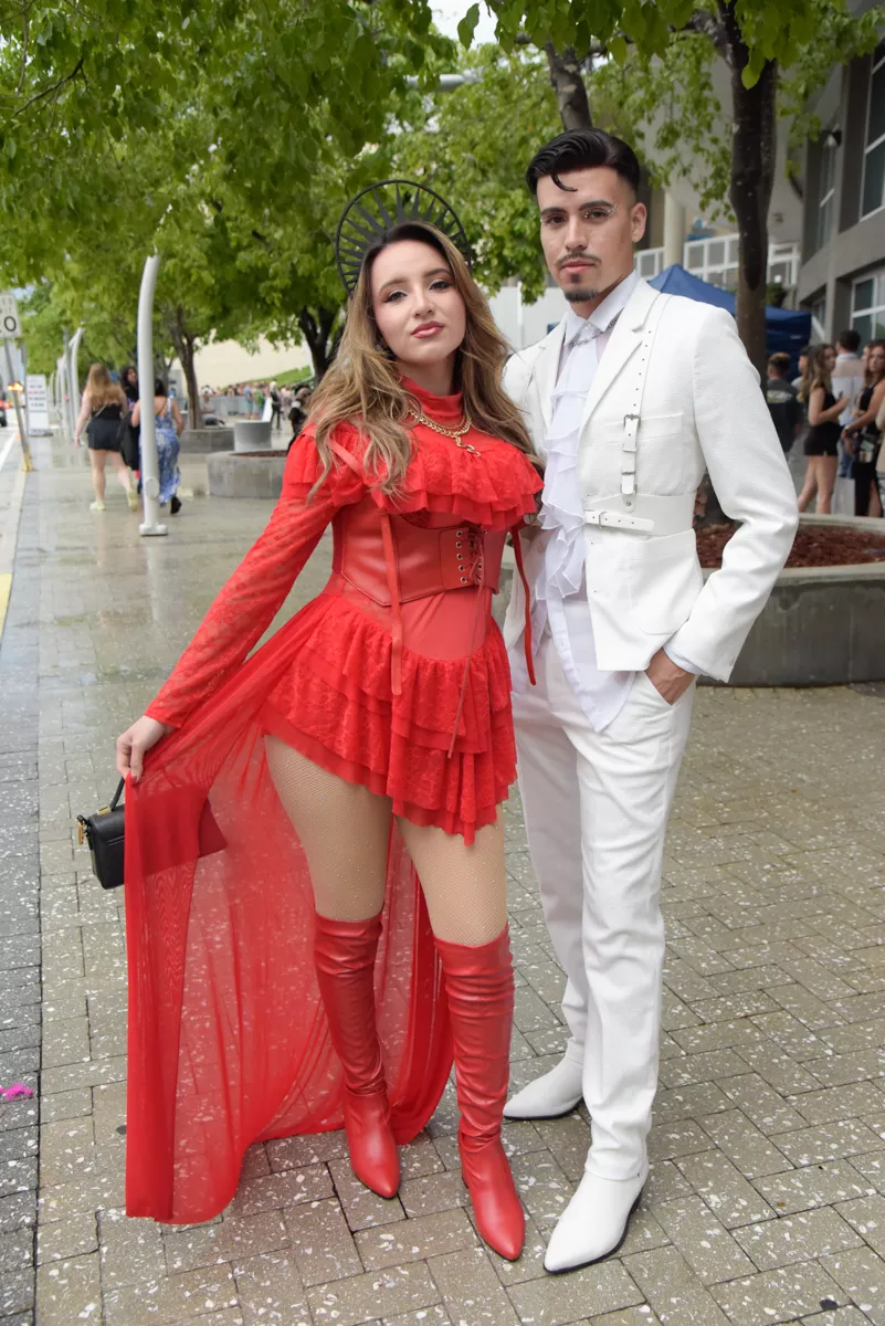 A woman wears a red lace, corseted dress with a black crown. The person next to her wears a white suit with a ruffled shirt