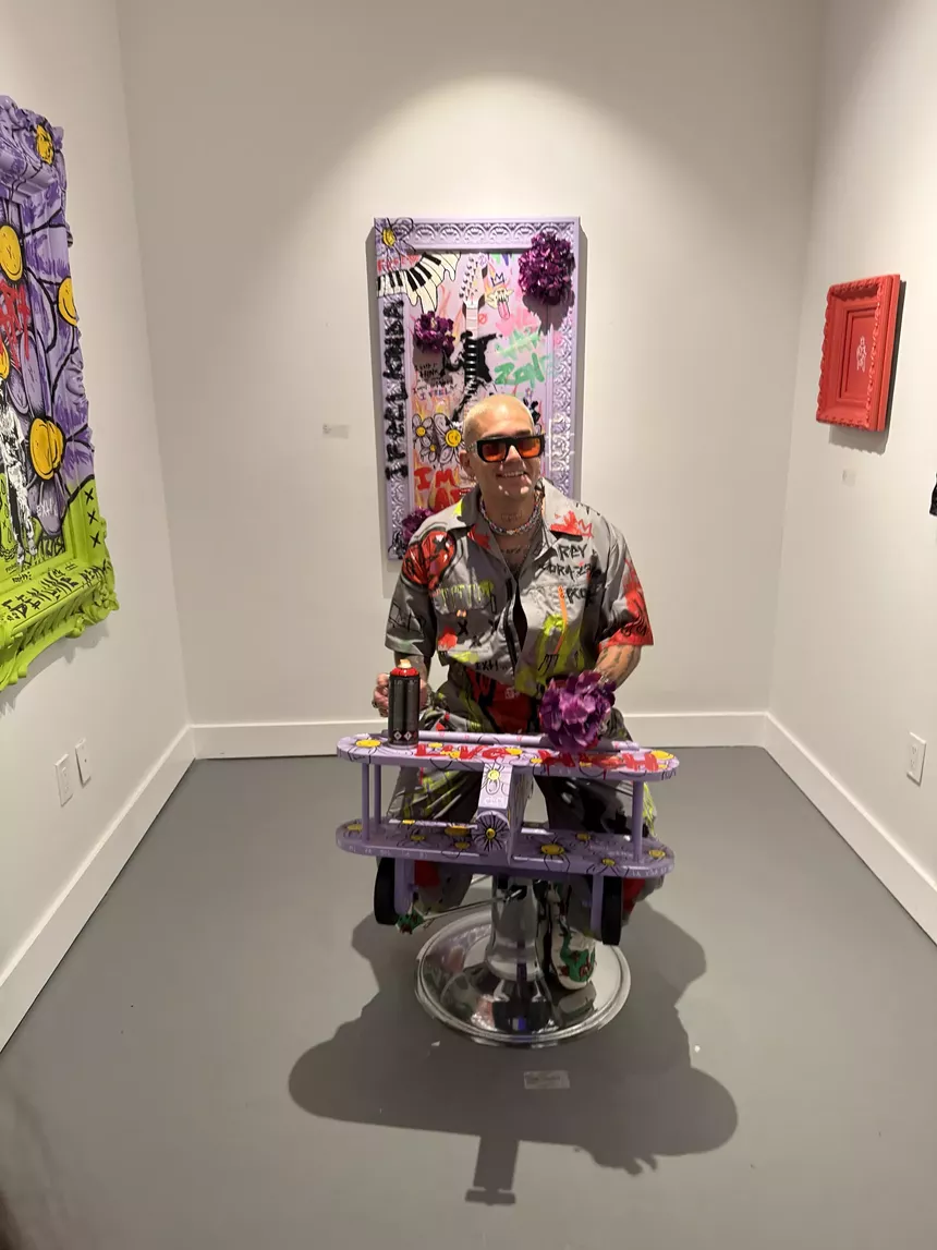 Picture of a man wearing colorful painter's overalls sitting in an art gallery.