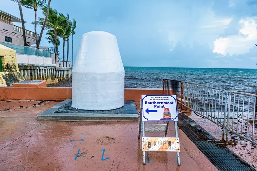 A white, concrete object shaped like a buoy in front of a seawall. A sign in front of it shows a painted replica of it reading "Southernmost Point," with a arrow pointing left