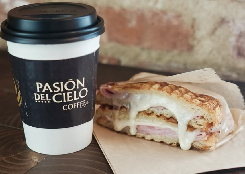 a grilled ham and cheese sandwich on a wood table next to a cup of coffee in a to-go cup