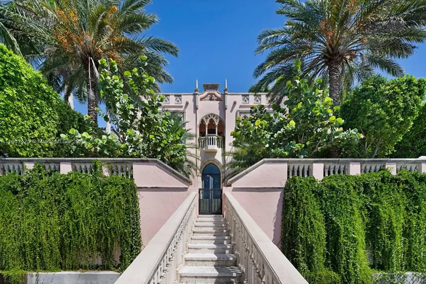 Depicted is a long outdoor walkway leading to a luxe front door of a Coconut Grove mansion.