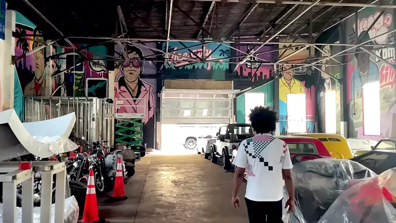 Image: Elo King walks through the interior of Vice City District, surrounded by colorful Miami Vice-inspired murals, vintage cars, and motorcycles in a large industrial warehouse space.