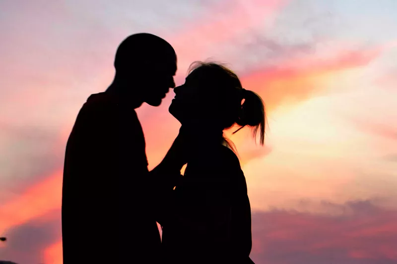 Image: A silhouetted couple leans in for a kiss with pink golden-hour skies behind them