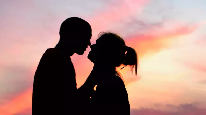 A silhouetted couple leans in for a kiss with pink golden-hour skies behind them