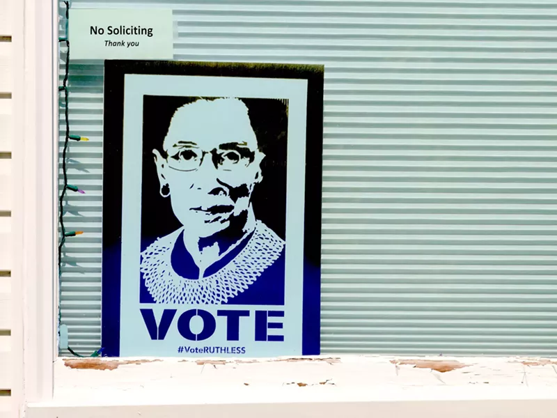 Image: small Justice Ruth Ginsburg "VOTE" poster displayed in a window underneath a smaller "no soliciting" sign