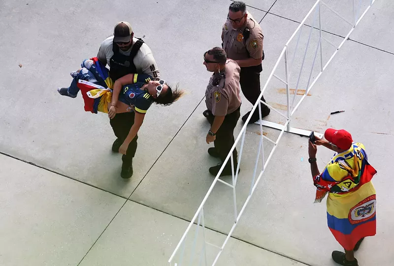 Image: uniformed police officer carrying a seemingly unconscious soccer outside a large sports stadium