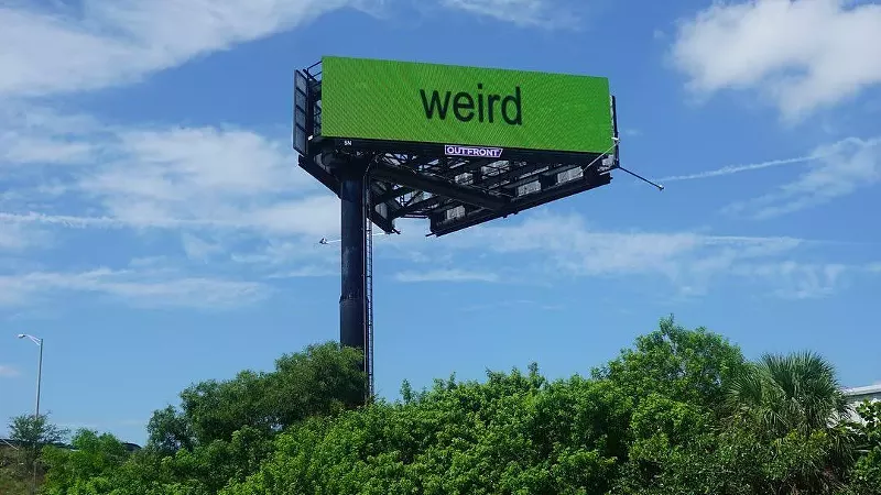 Image: billboard with the word "weird" spelled out in lowercase sans serif letters against a bright green background, set against a verdant South Florida landscape on a bright sunny day