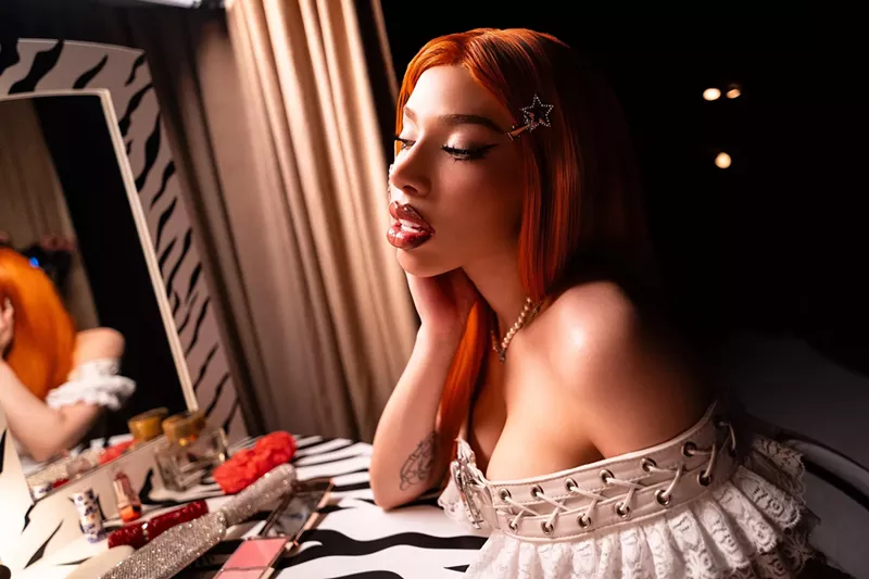 Image: A heavily made-up woman with long red hair, seated at a dressing table