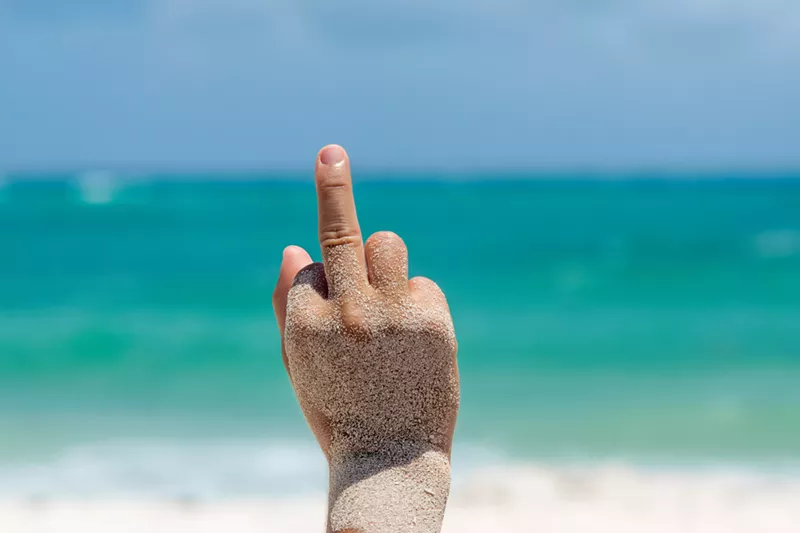Image: Female hand covered with sand performs a Fuck you Sign with her hand in a tropical beach